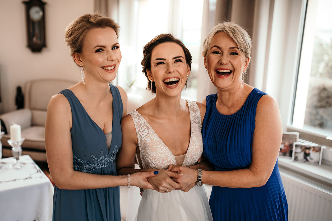 Panna młoda w białej sukni ślubnej, trzymająca dłonie z dwiema uśmiechniętymi kobietami w niebieskich sukienkach, śmieją się radośnie w jasnym wnętrzu.