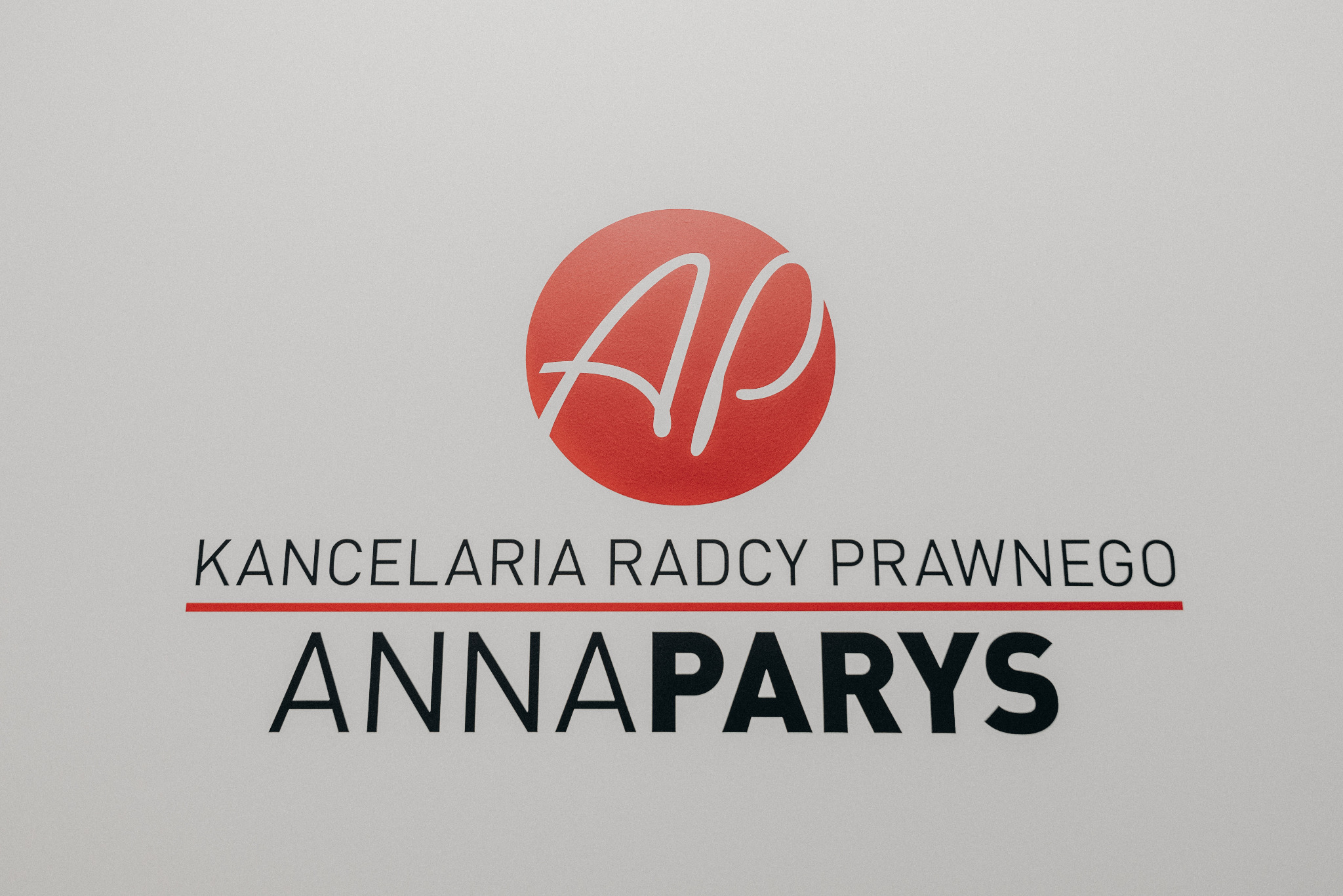 Logo kancelarii prawnej Anna Parys: czerwony okrąg z białymi inicjałami AP, poniżej napis Kancelaria Radcy Prawnego i imię i nazwisko Anna Parys, wszystko na jasnoszarym tle.