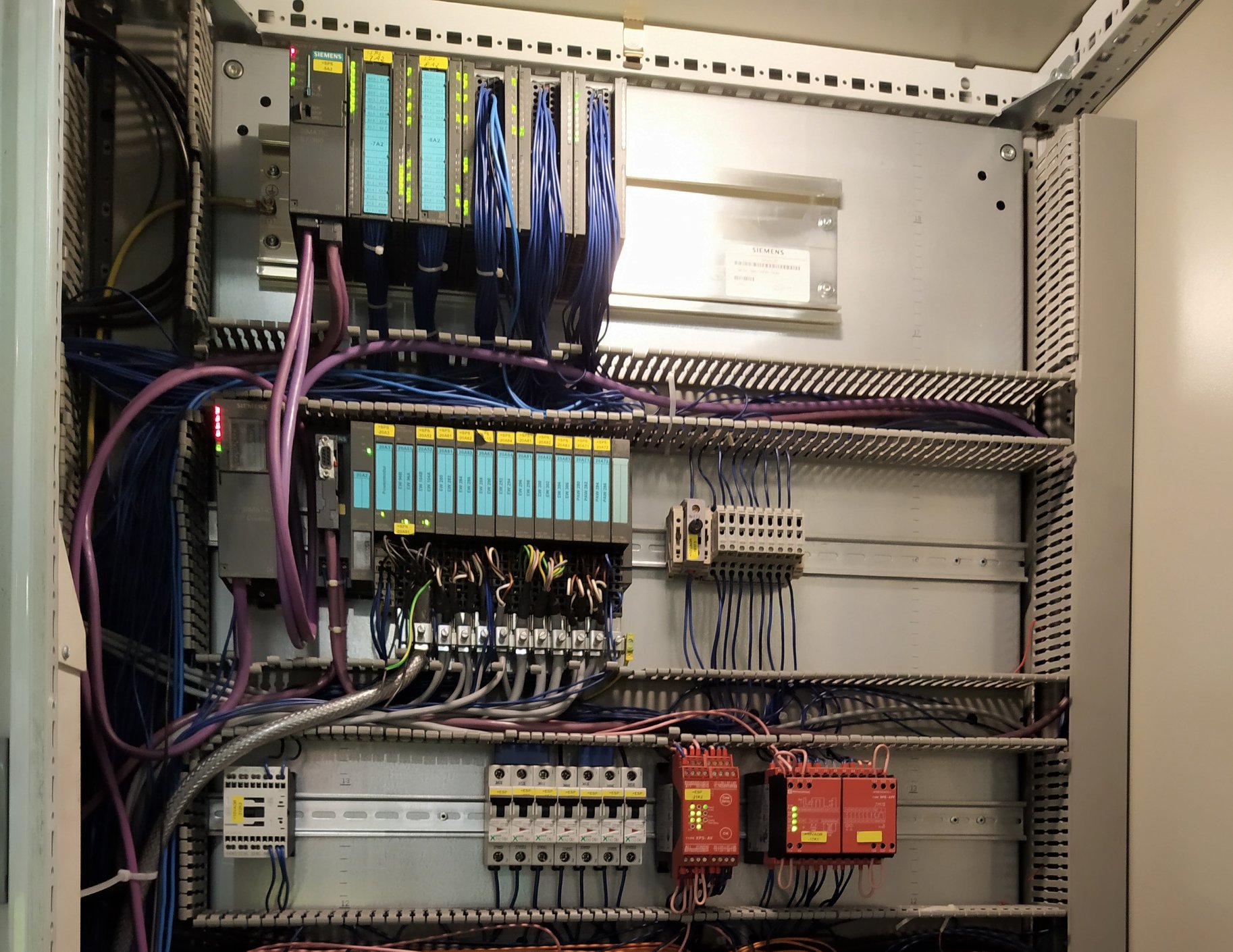 Wnętrze szafy sterowniczej z modułami PLC Siemens, okablowaniem w kolorach niebieskim i fioletowym, oraz przekaźnikami bezpieczeństwa w obudowach koloru czerwonego.