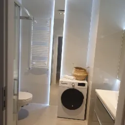 Nowoczesna łazienka w odcieniach szarości z pralką, kabiną prysznicową i podświetlanym lustrem, uzupełniona o dekoracyjny koszyk na pralce.