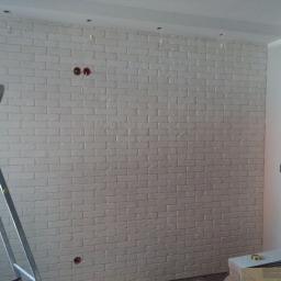 Wikrem - Ściana wyłożona białą cegłą dekoracyjną, widoczne puszki elektryczne bez osprzętu, drabina aluminiowa i stos płytek pod ścianą.