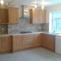 Wikrem - Jasna kuchnia z drewnianymi szafkami, mozaikową ścianą nad blatem, stalowym okapem i białymi płytkami podłogowymi, widok na zewnątrz przez okno.