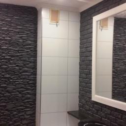 Wikrem - Wykończona łazienka z akcentami: ściana z szarego kamienia dekoracyjnego, fragment ściany z białych płytek i ściana z czarnej cegły, biała rama lustra i ciemne drzwi.