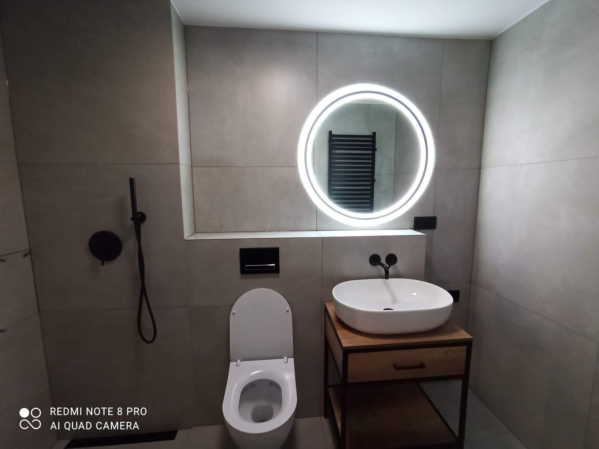 Nowoczesna łazienka z szarymi płytkami, okrągłym lustrem z podświetleniem LED, wiszącą umywalką na drewnianej szafce, wiszącą toaletą i czarną armaturą.