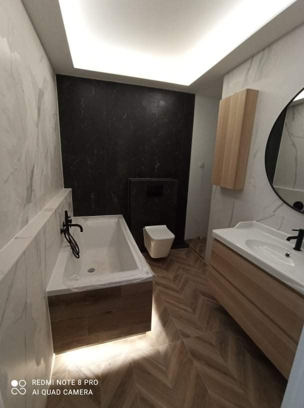 Nowoczesna łazienka po remoncie w Szewcach, z marmurowymi płytkami na ścianach, czarną ścianą akcentującą, podświetlaną wanną i drewnianą szafką pod umywalką oraz podłogą ułożoną w jodełkę.