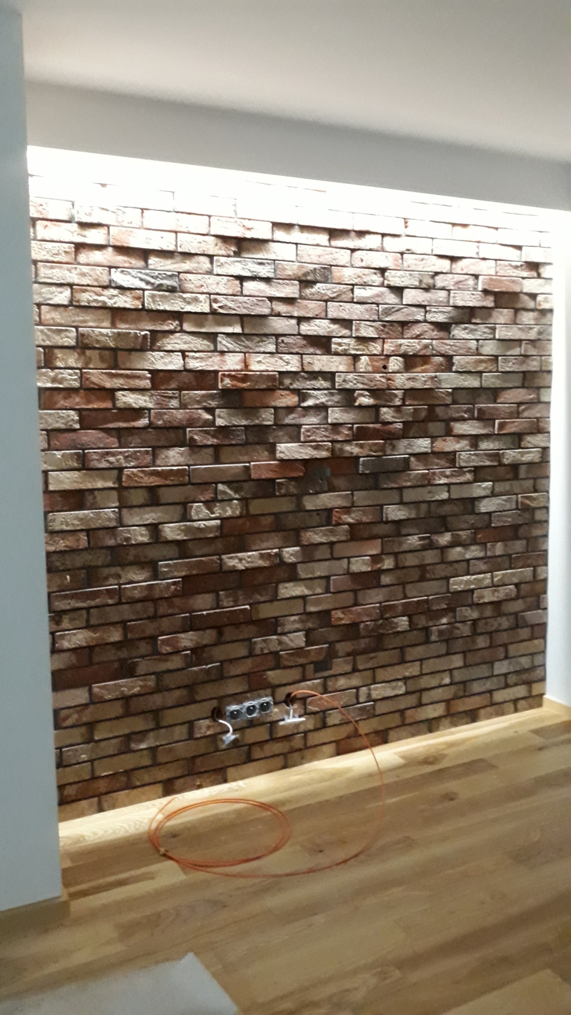Ściana wykończona dekoracyjną cegłą w odcieniach brązu i beżu, z widocznymi gniazdkami elektrycznymi i fragmentem podłogi z paneli drewnopodobnych, oświetlona listwą LED w górnej części.