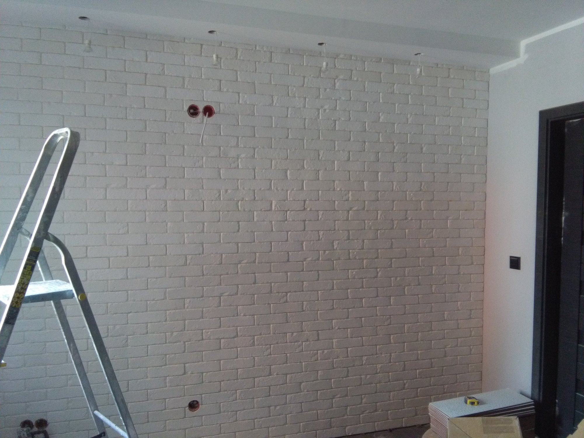 Ściana wyłożona białą cegłą dekoracyjną, widoczne puszki elektryczne bez osprzętu, drabina aluminiowa i stos płytek pod ścianą.