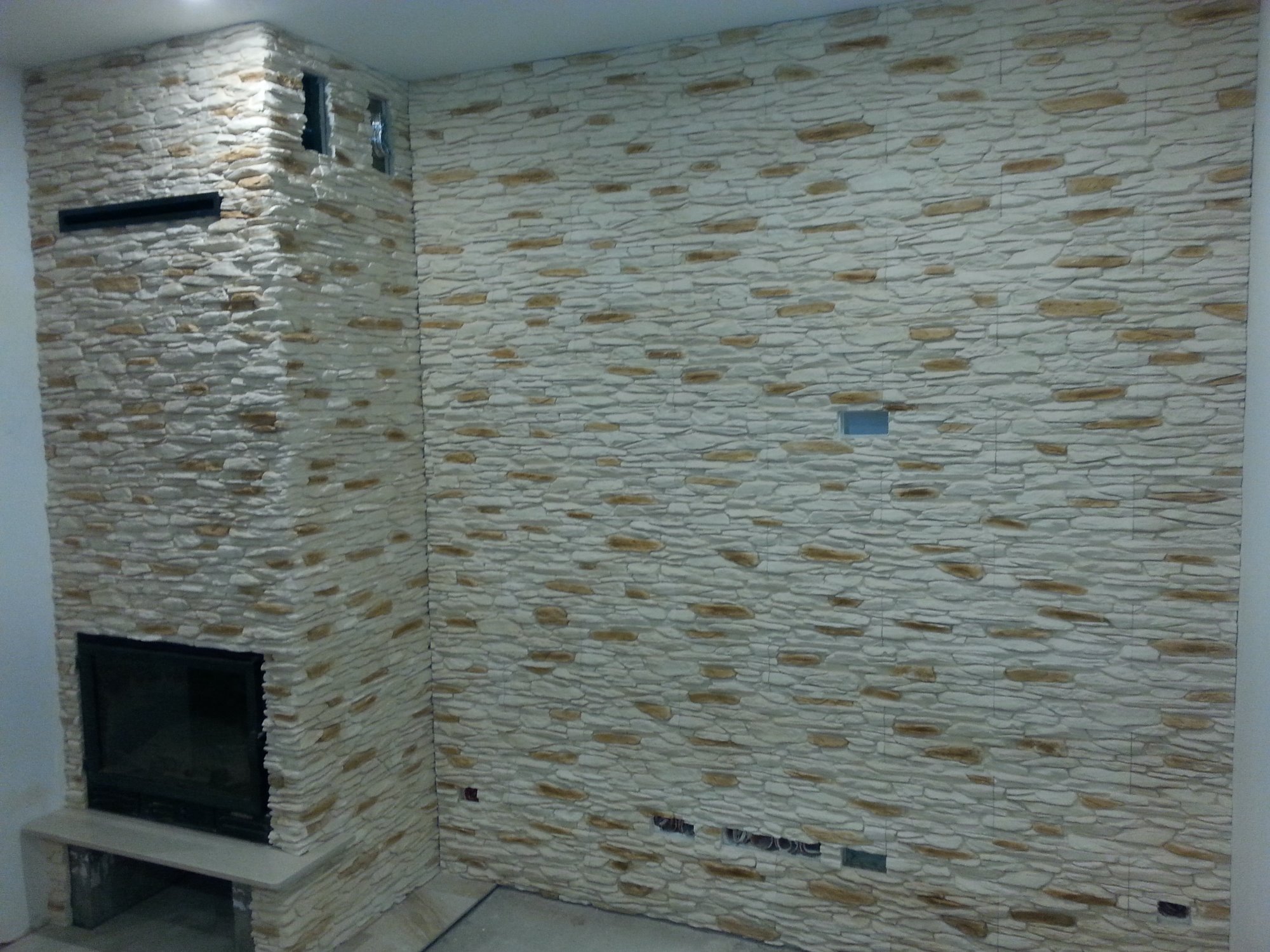 Ściana wykończona dekoracyjnym kamieniem z imitacją piaskowca, zabudowa kominka z czarnym wkładem, widoczne otwory na gniazdka elektryczne w ścianie.