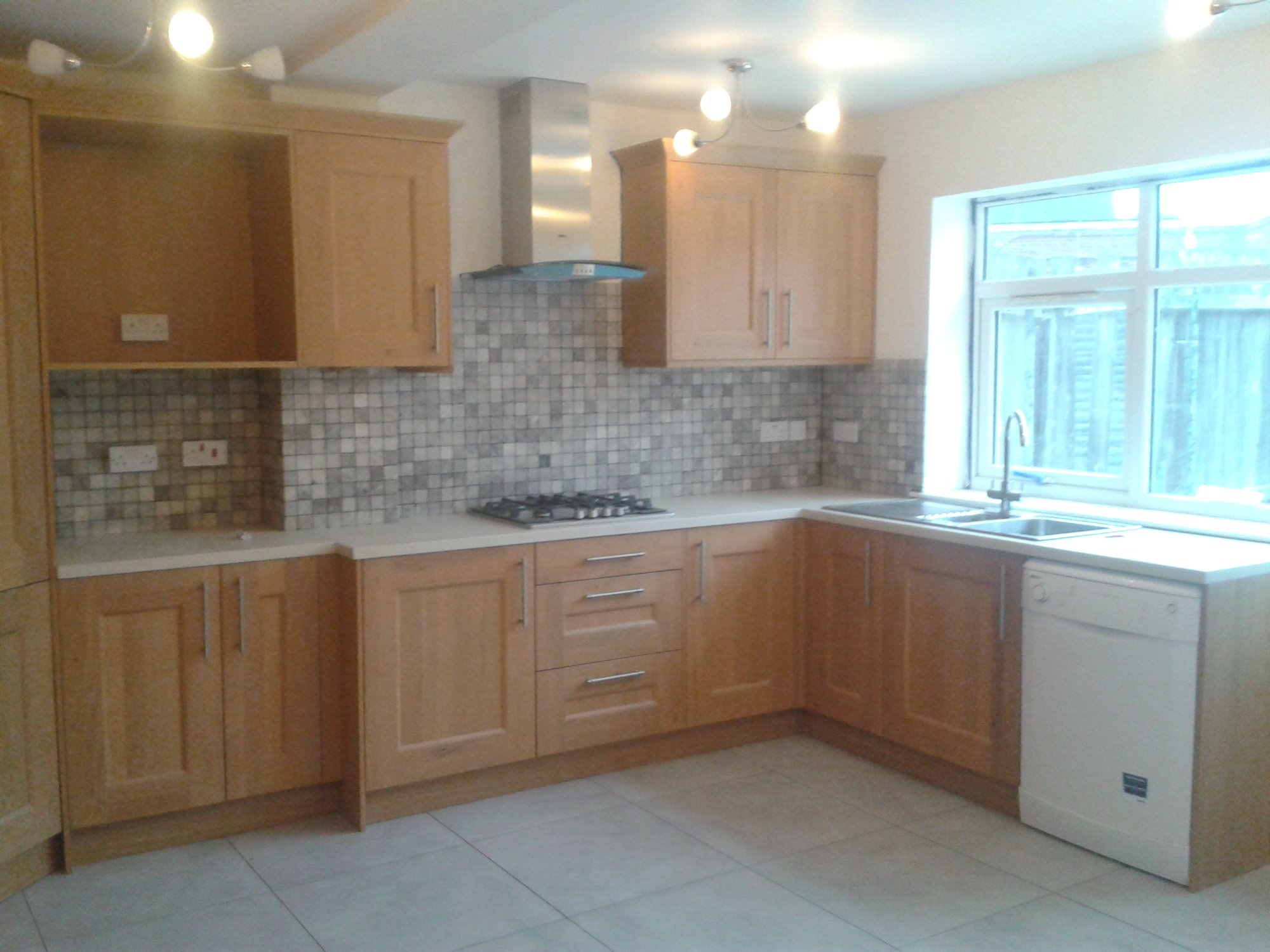 Jasna kuchnia z drewnianymi szafkami, mozaikową ścianą nad blatem, stalowym okapem i białymi płytkami podłogowymi, widok na zewnątrz przez okno.