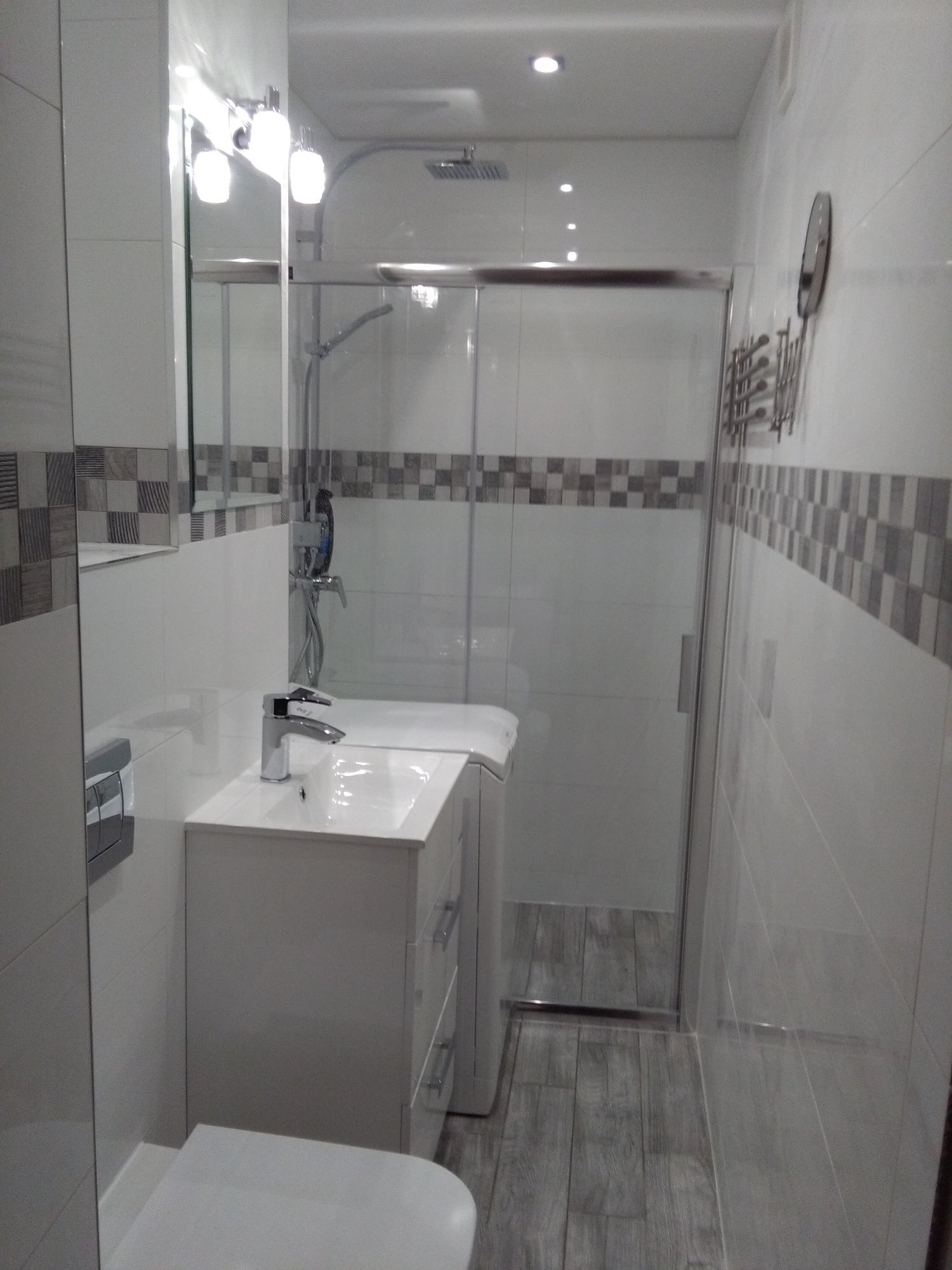 Jasna łazienka z umywalką nablatową, kabiną prysznicową ze szklanymi drzwiami przesuwnymi, lustrem z oświetleniem punktowym i dekoracyjnym pasem płytek w szaro-białą szachownicę.