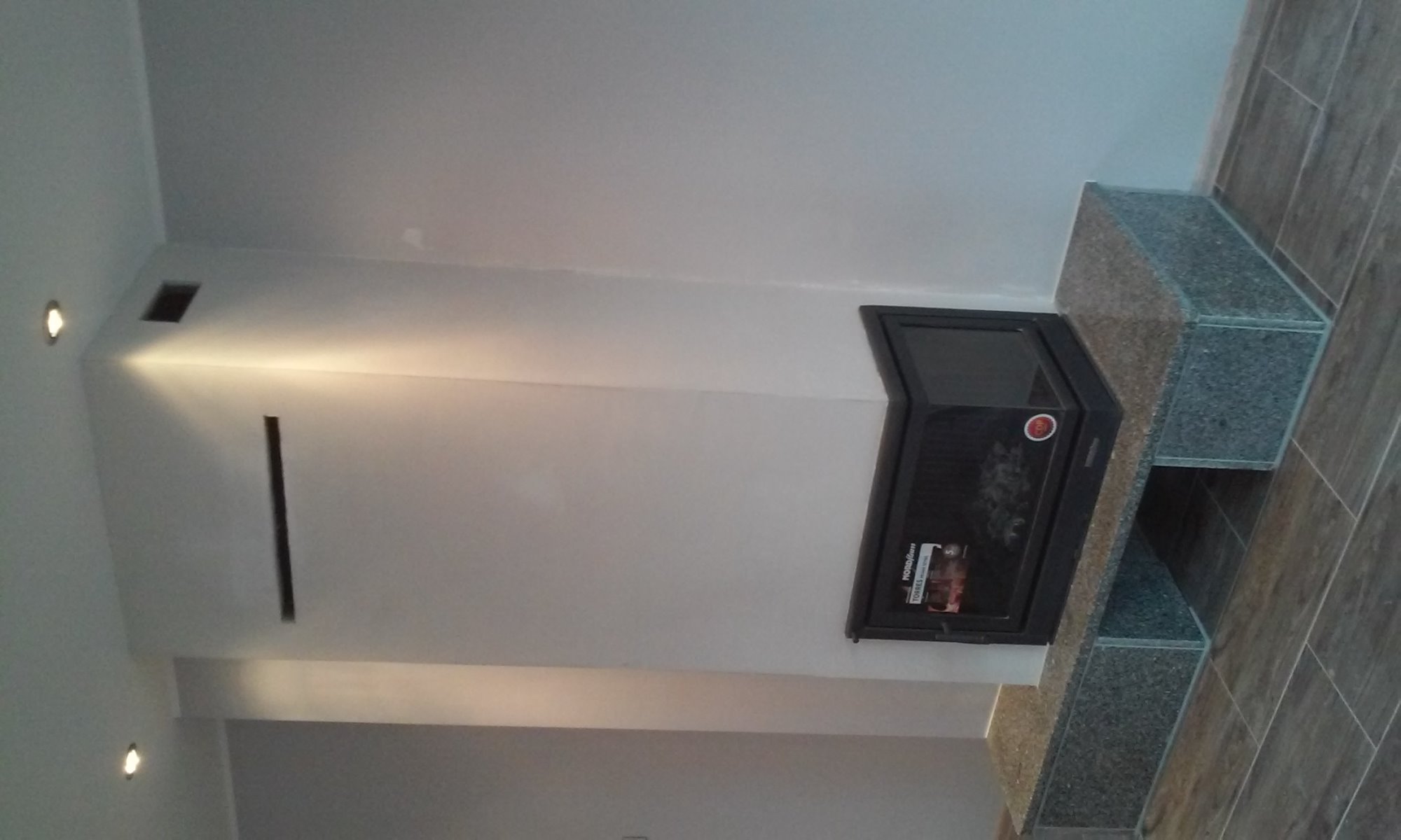 Nowoczesny kominek narożny z czarną obudową i granitową zabudową, oświetlenie punktowe w suficie, widok z dołu.