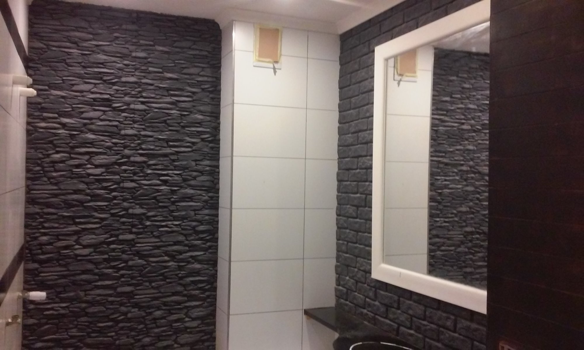 Wykończona łazienka z akcentami: ściana z szarego kamienia dekoracyjnego, fragment ściany z białych płytek i ściana z czarnej cegły, biała rama lustra i ciemne drzwi.