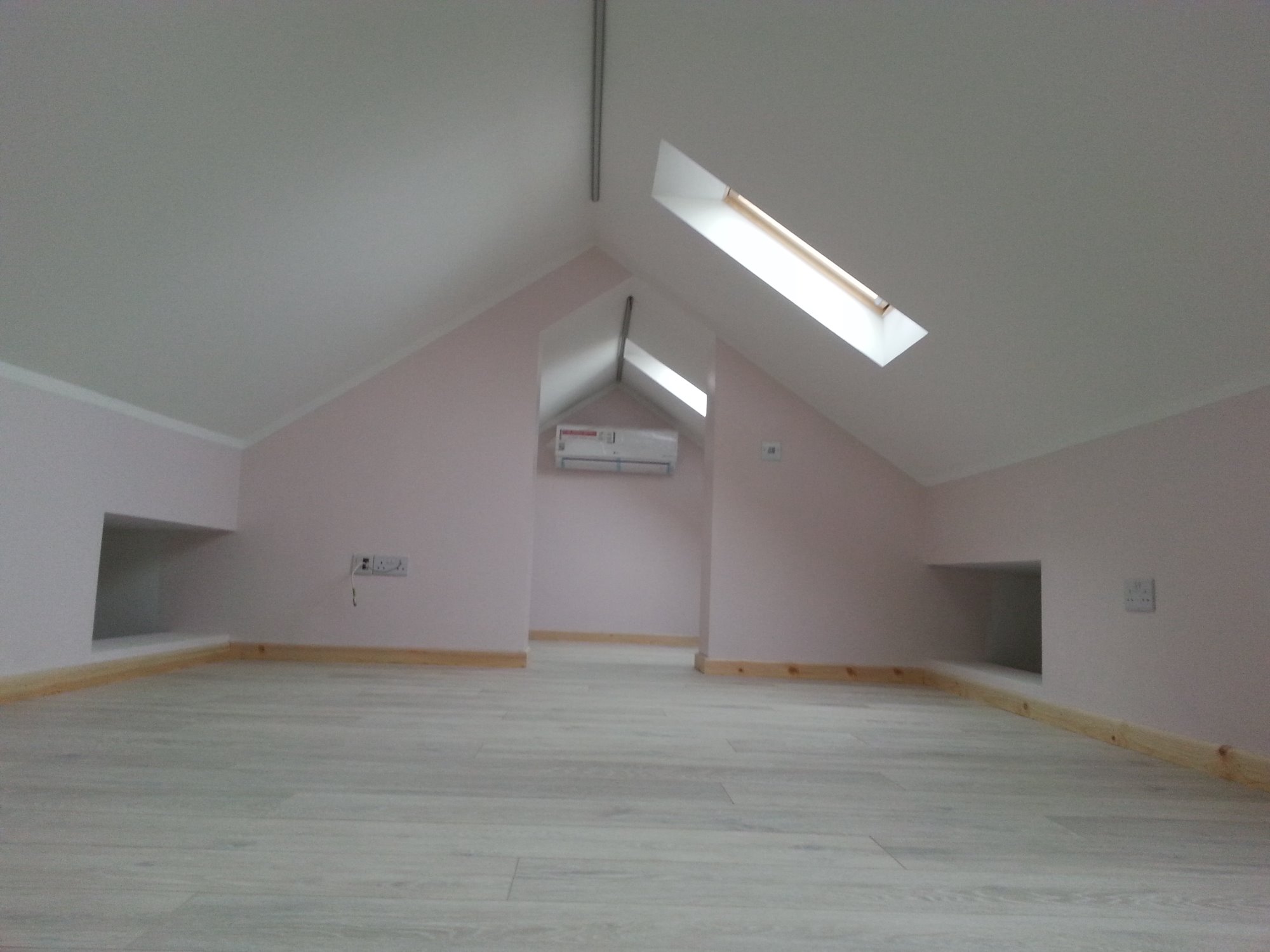 Poddasze po remoncie z jasnymi panelami, różowymi ścianami, oknem dachowym i wnękami w ścianach.