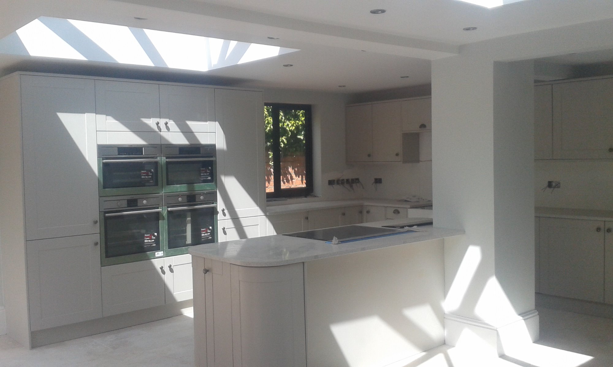 Jasna kuchnia w trakcie remontu, z czterema piekarnikami w zabudowie, wyspą kuchenną i oknem dachowym wpuszczającym promienie słoneczne.