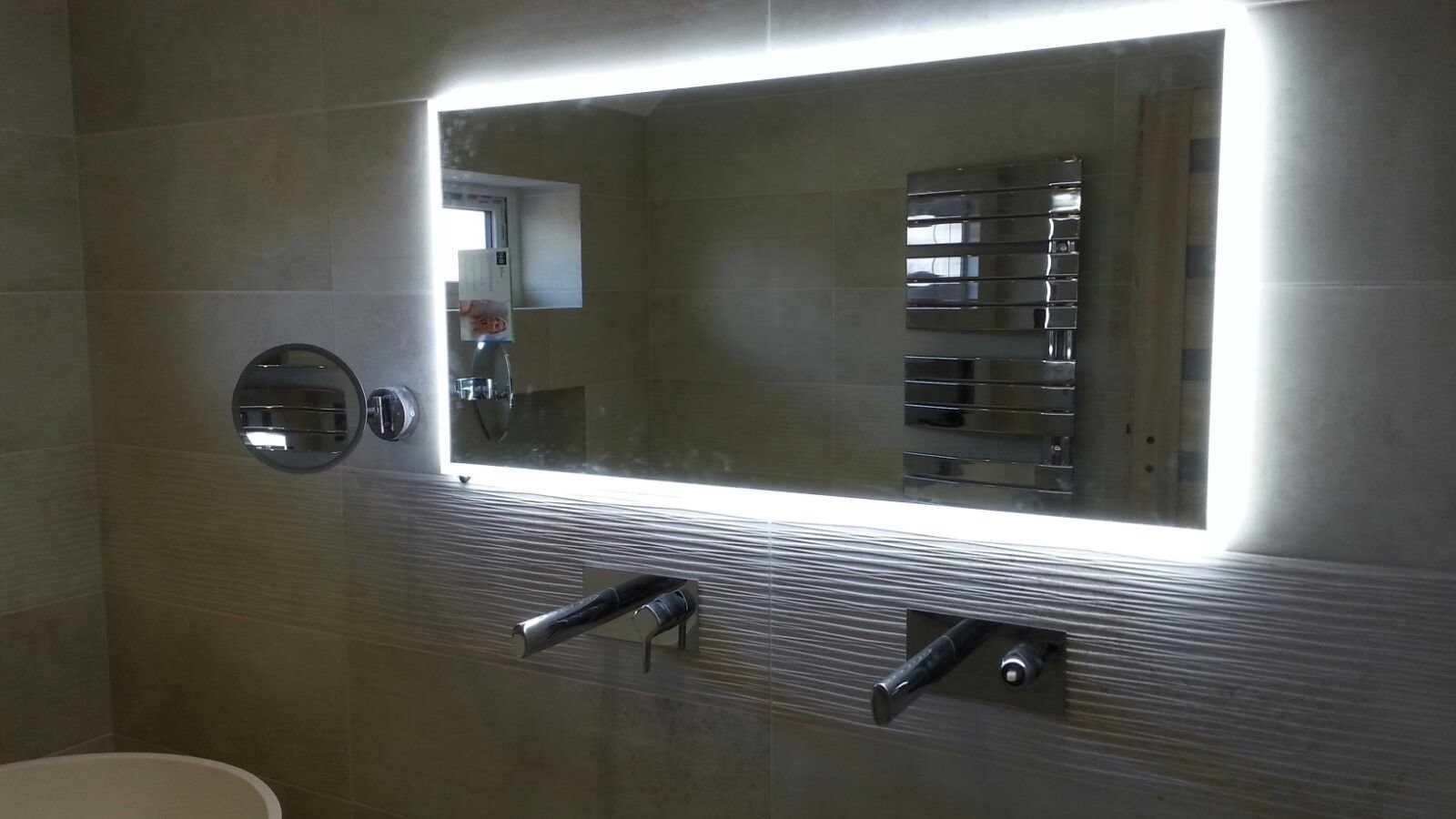 Nowoczesna łazienka w Szewcach po remoncie: podświetlane lustro, armatura podtynkowa i grzejnik drabinkowy na tle beżowych płytek z delikatną fakturą.