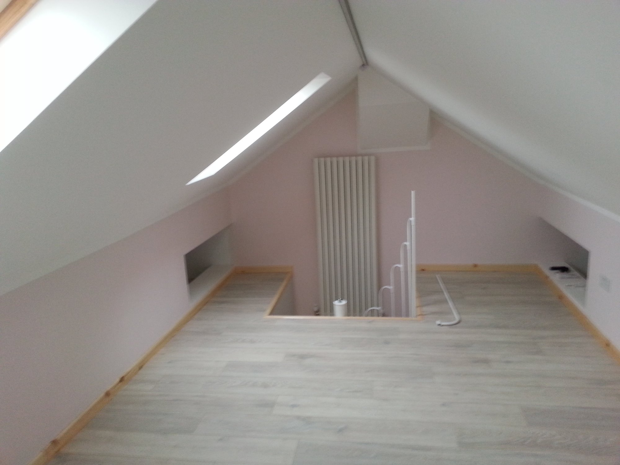 Poddasze po remoncie z jasnymi panelami podłogowymi, grzejnikiem i wnękami w ścianach, wykończone drewnianymi listwami przypodłogowymi.