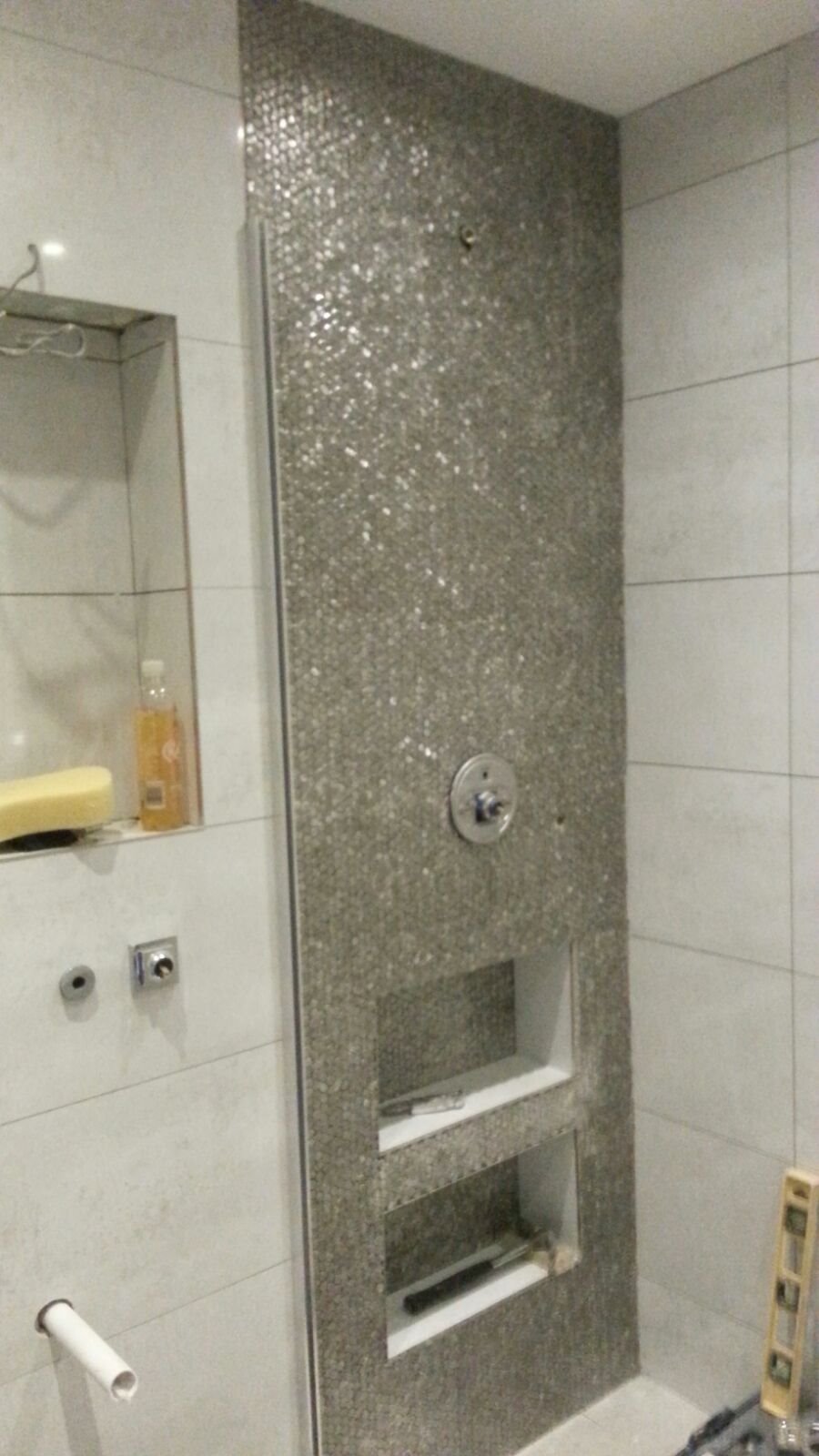 Ściana prysznicowa wyłożona mozaiką z wnękami na kosmetyki, widoczny zawór sterujący i fragment poziomicy na podłodze, w trakcie prac wykończeniowych.