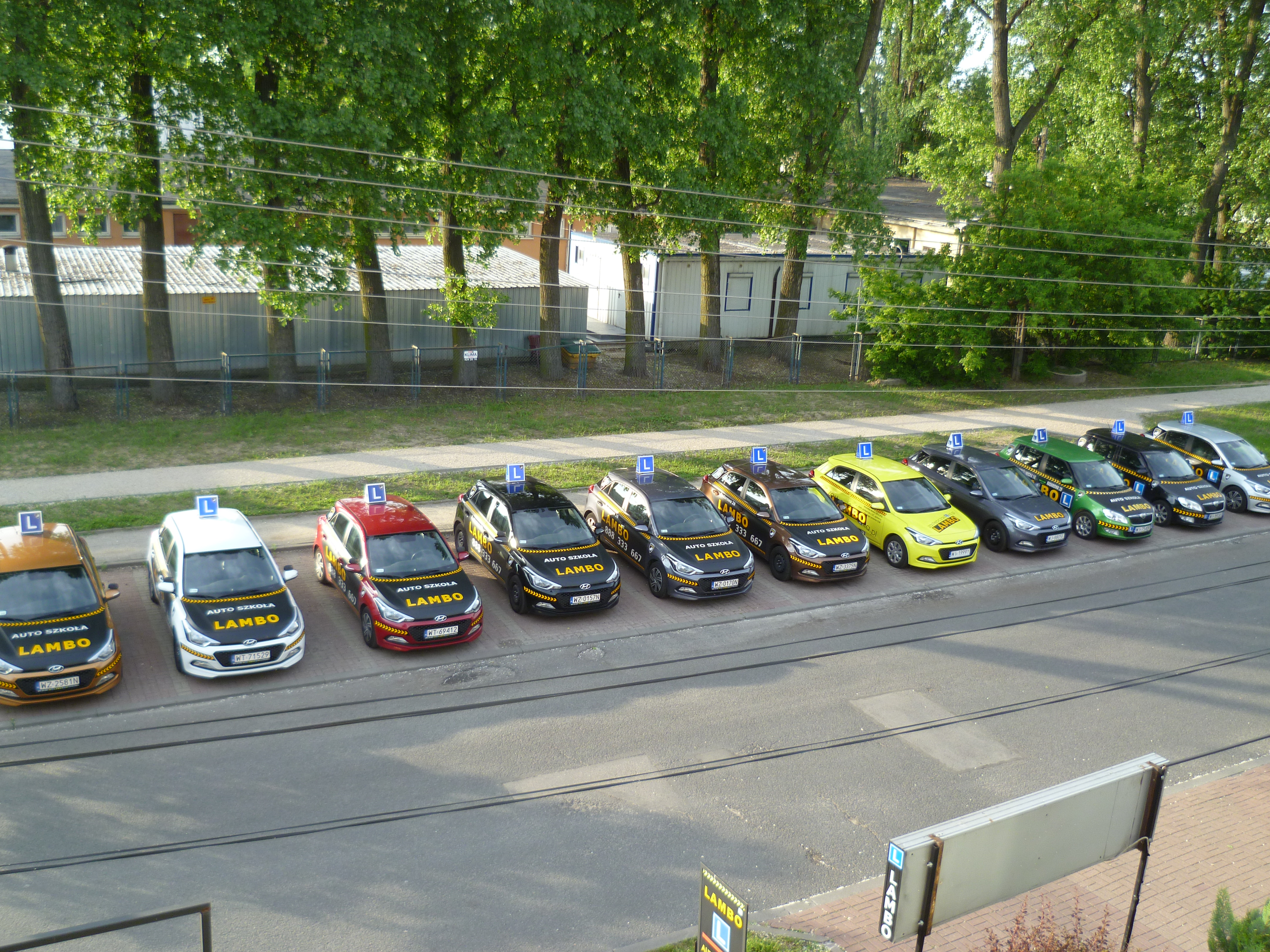 Flota samochodów o różnych kolorach z naklejkami 'Auto Szkoła Lambo' i znakiem 'L' na dachu, zaparkowana w rzędzie przed budynkiem i drzewami.