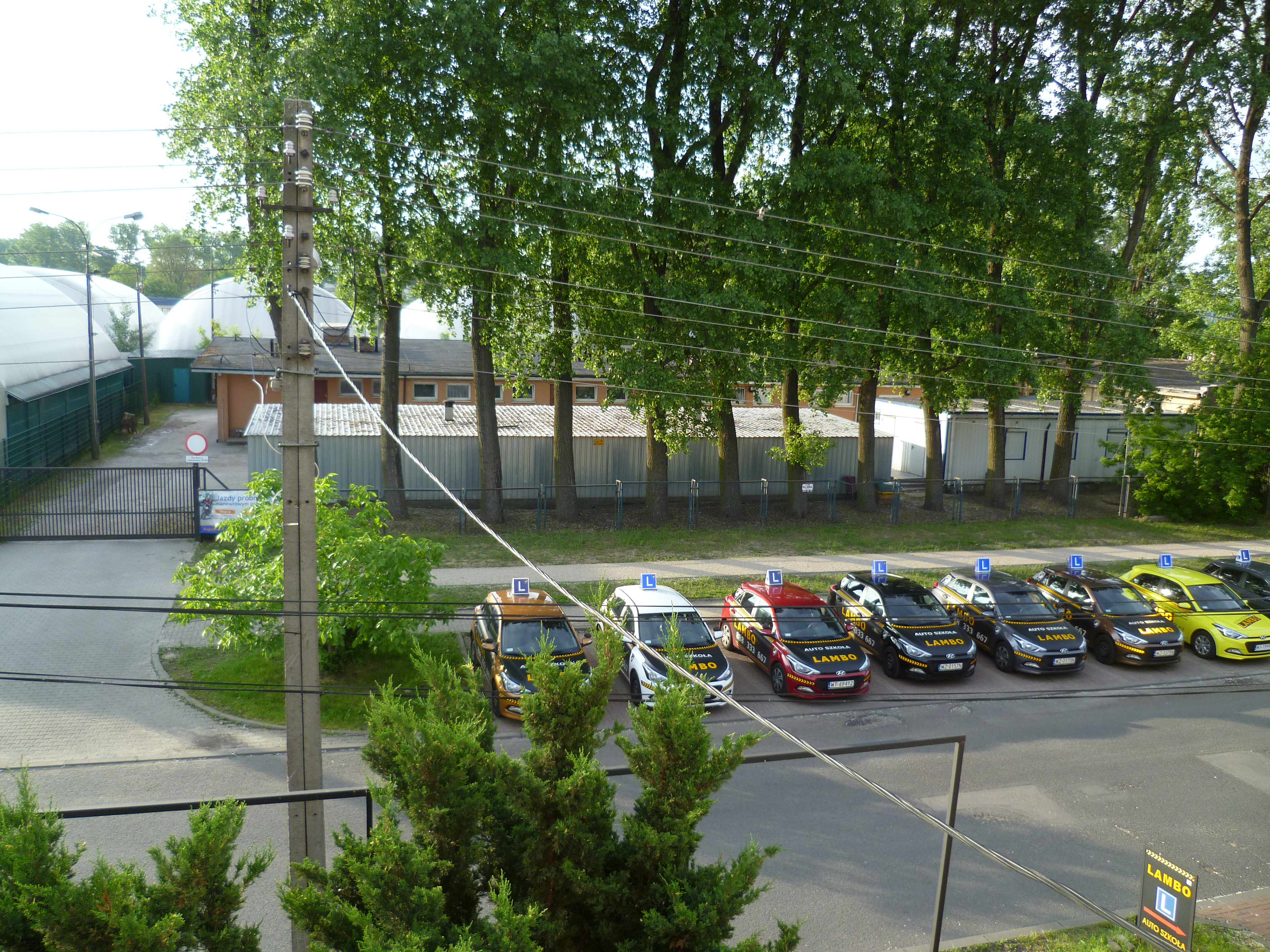 Flota samochodów o różnych kolorach z naklejkami z nazwą firmy zaparkowana przed budynkiem na tle zieleni i słupa energetycznego.