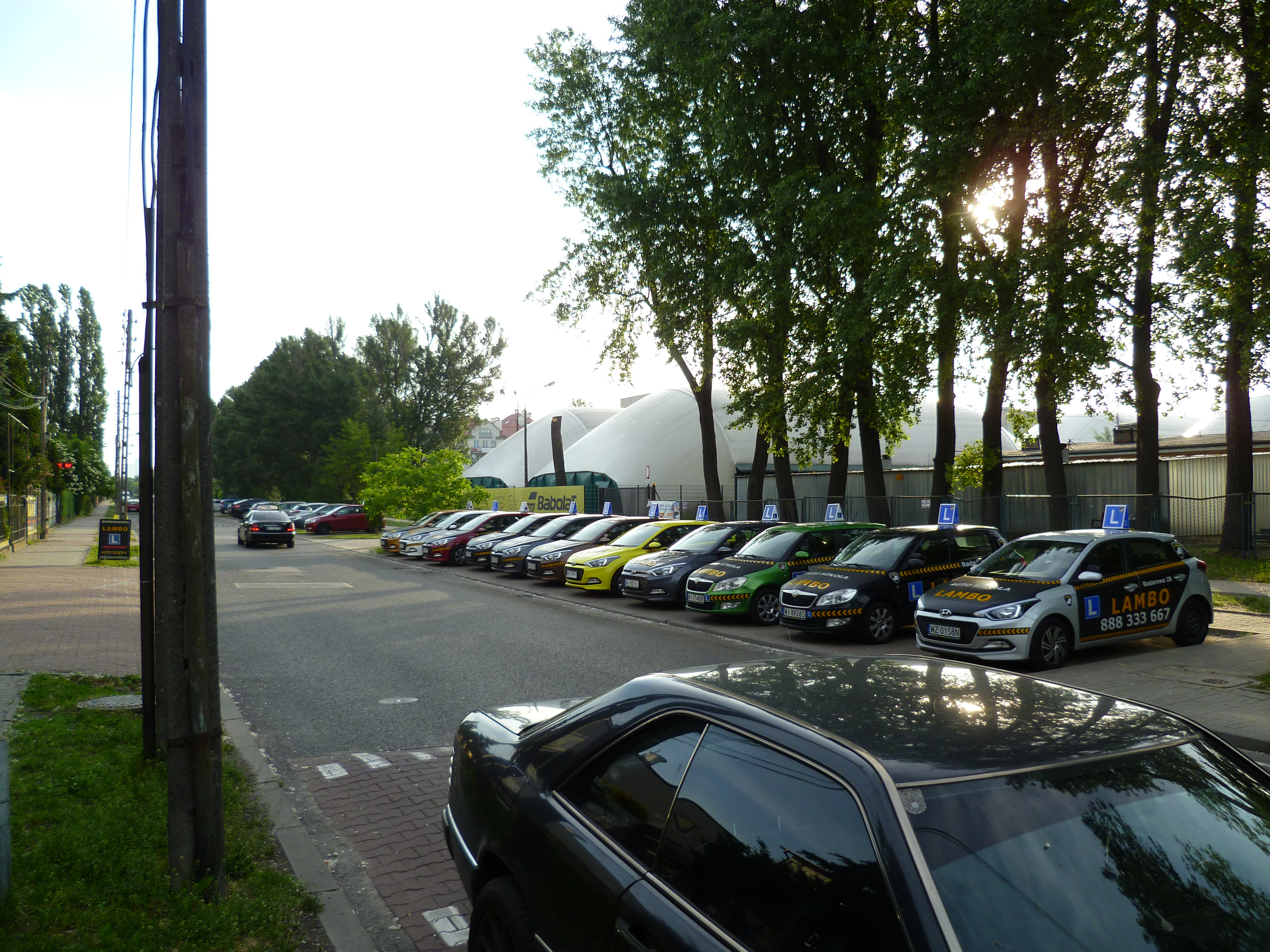 Rząd samochodów oznaczonych literą 'L' zaparkowanych wzdłuż ulicy, z budynkiem o białej kopule w tle i widocznym znakiem Auto Szkoła.