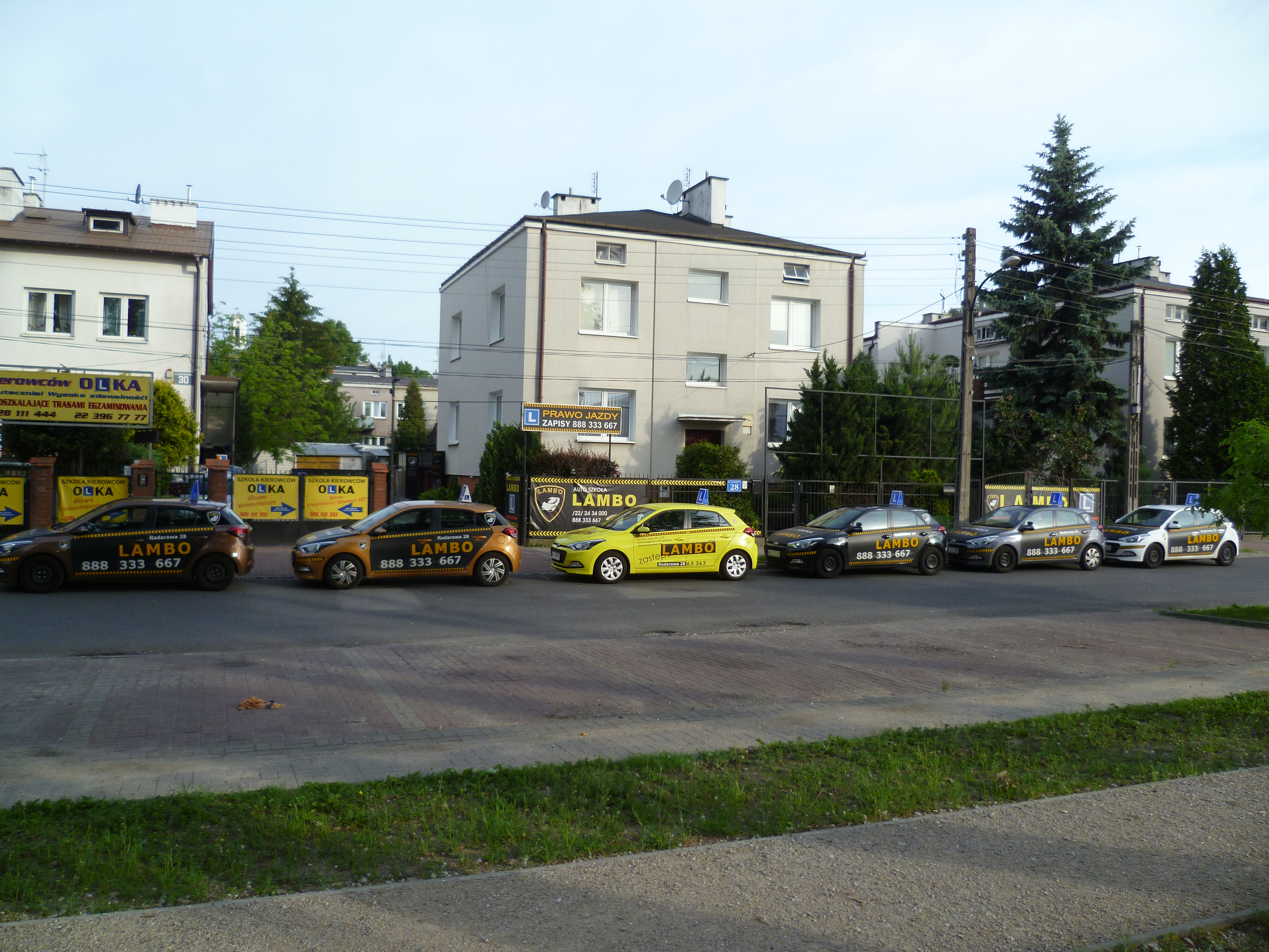 Rząd samochodów oznaczonych logo i numerem telefonu, zaparkowanych przed budynkami, prawdopodobnie należących do firmy oferującej kursy prawa jazdy.