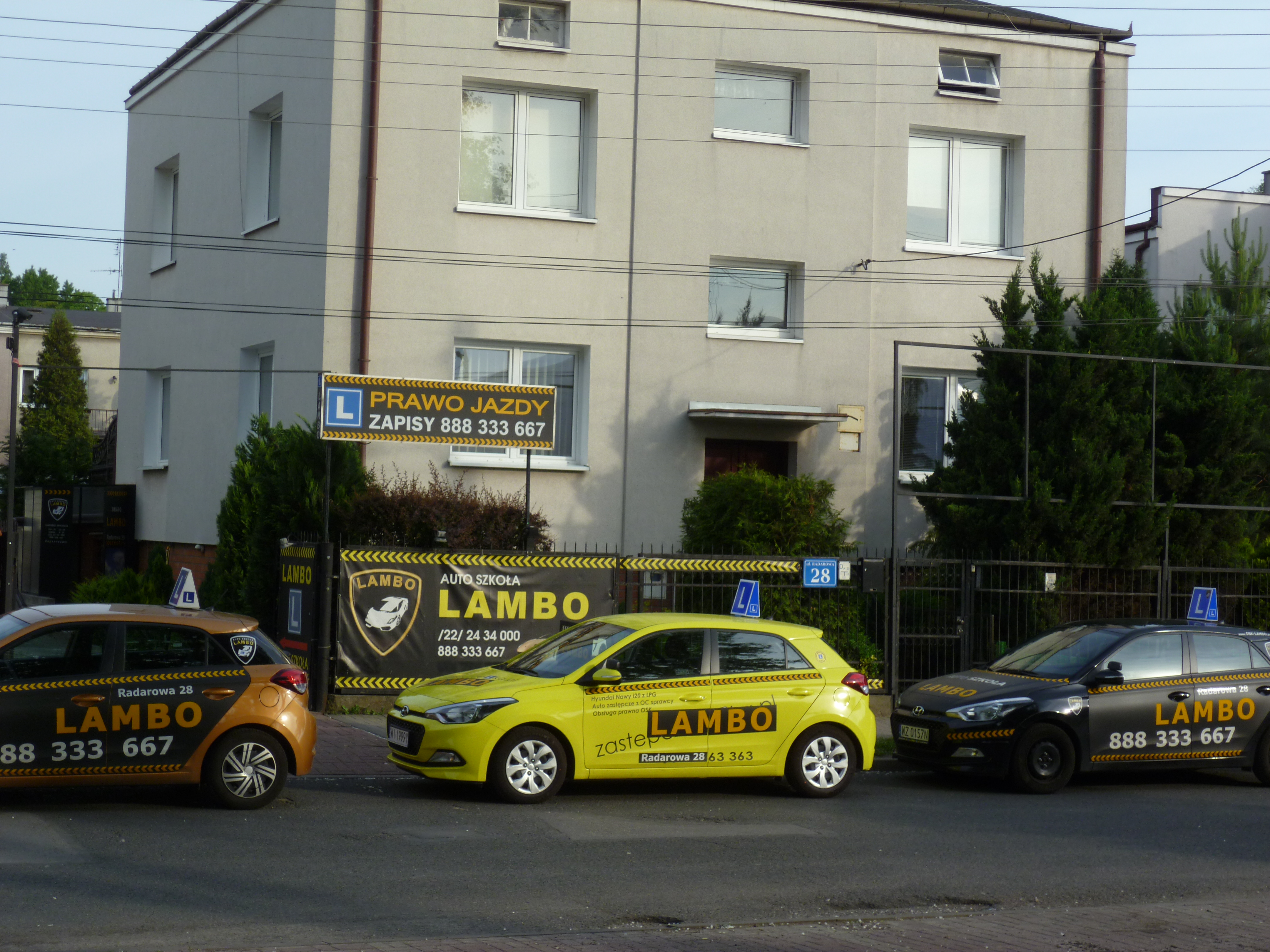 Flota samochodów oznaczonych logo szkoły jazdy LAMBO zaparkowana przed budynkiem szkoły z szyldem 'Prawo Jazdy, Zapisy 888 333 667'.
