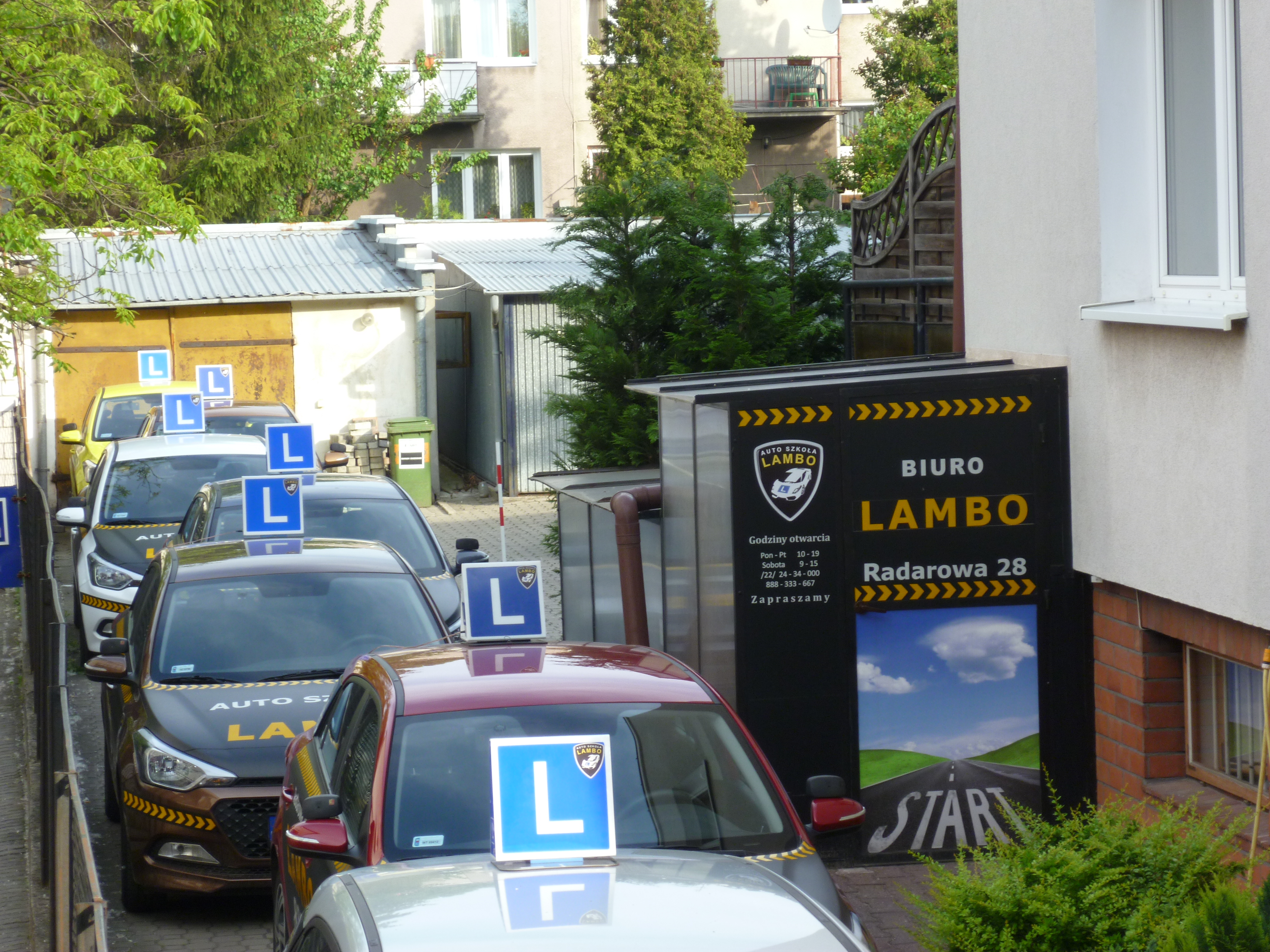 Flota samochodów oznaczonych literą 'L' na dachu, należących do Auto Szkoły Lambo, zaparkowana przed budynkiem z reklamą szkoły jazdy.