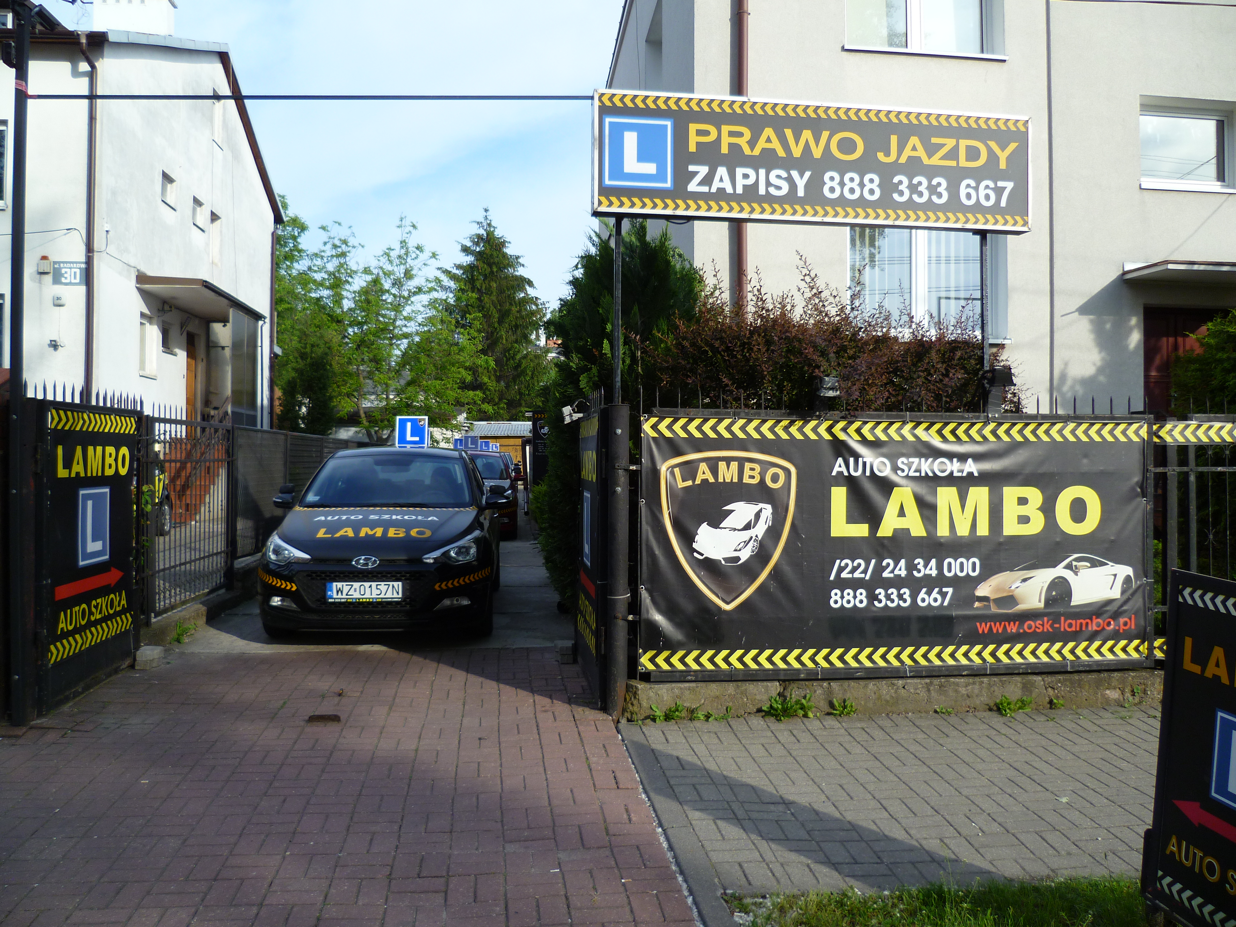 Wjazd do auto szkoły z zaparkowanymi samochodami oznaczonymi logo firmy, widoczny szyld z informacją o zapisach i numerem telefonu.