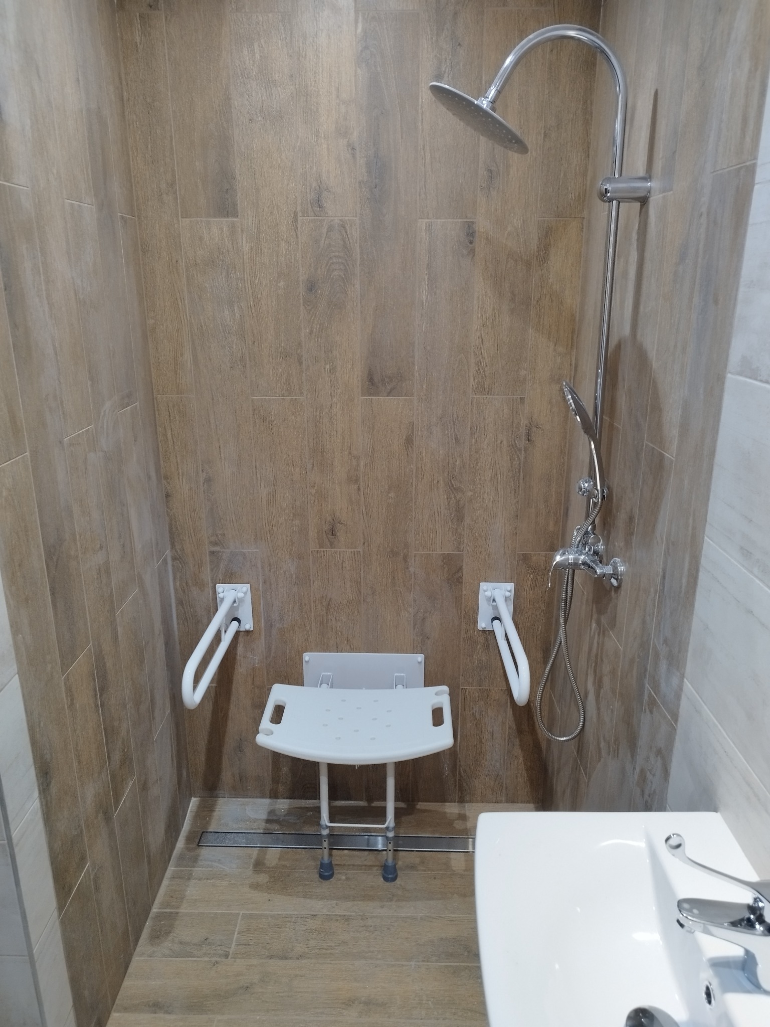 Kabina prysznicowa z siedziskiem i uchwytami dla osób z niepełnosprawnościami, wykończona płytkami imitującymi drewno, z widoczną umywalką po prawej stronie.