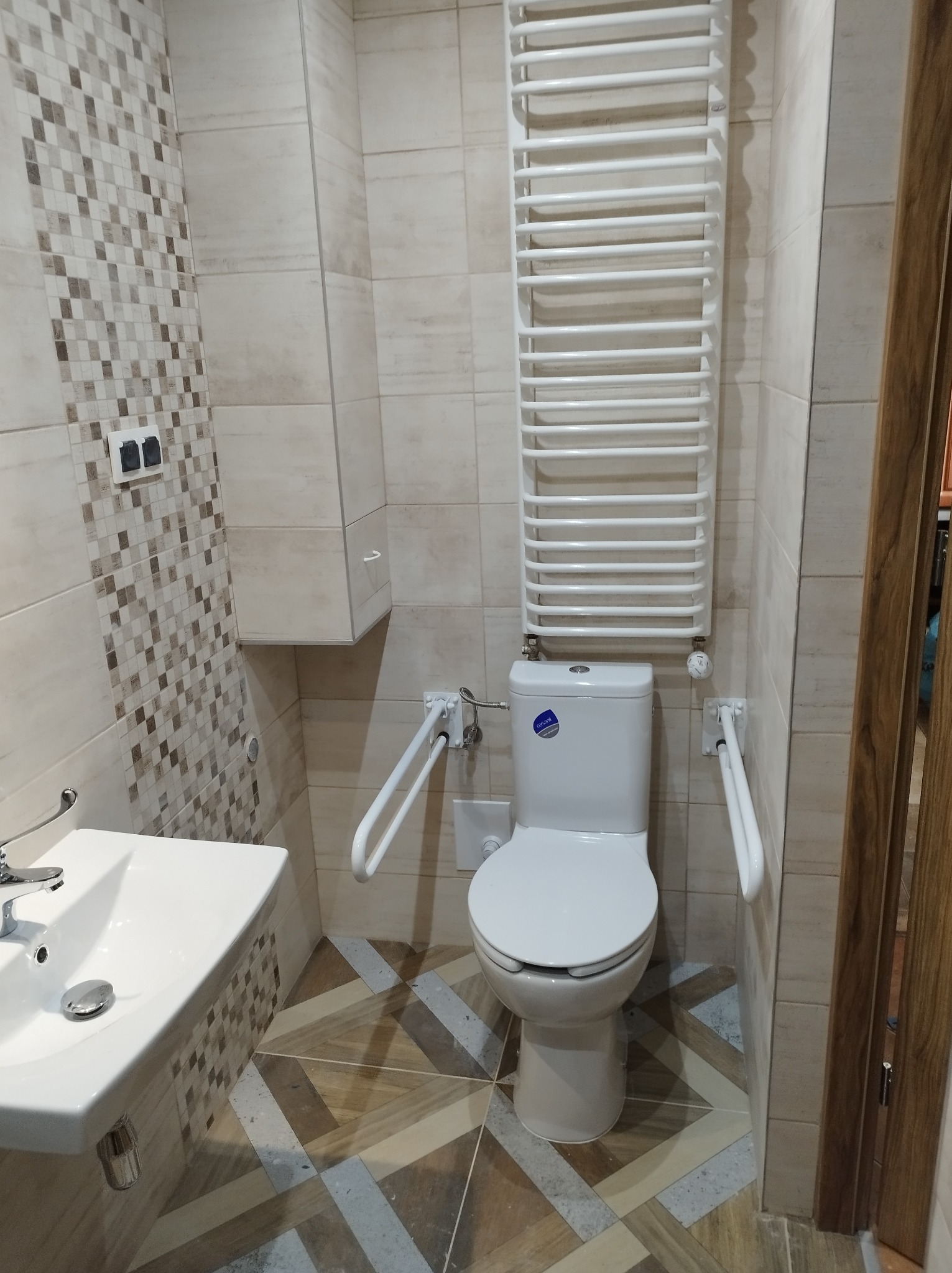 Wykończona łazienka z toaletą z uchwytami dla niepełnosprawnych, grzejnikiem drabinkowym, umywalką i mozaikową ścianą.