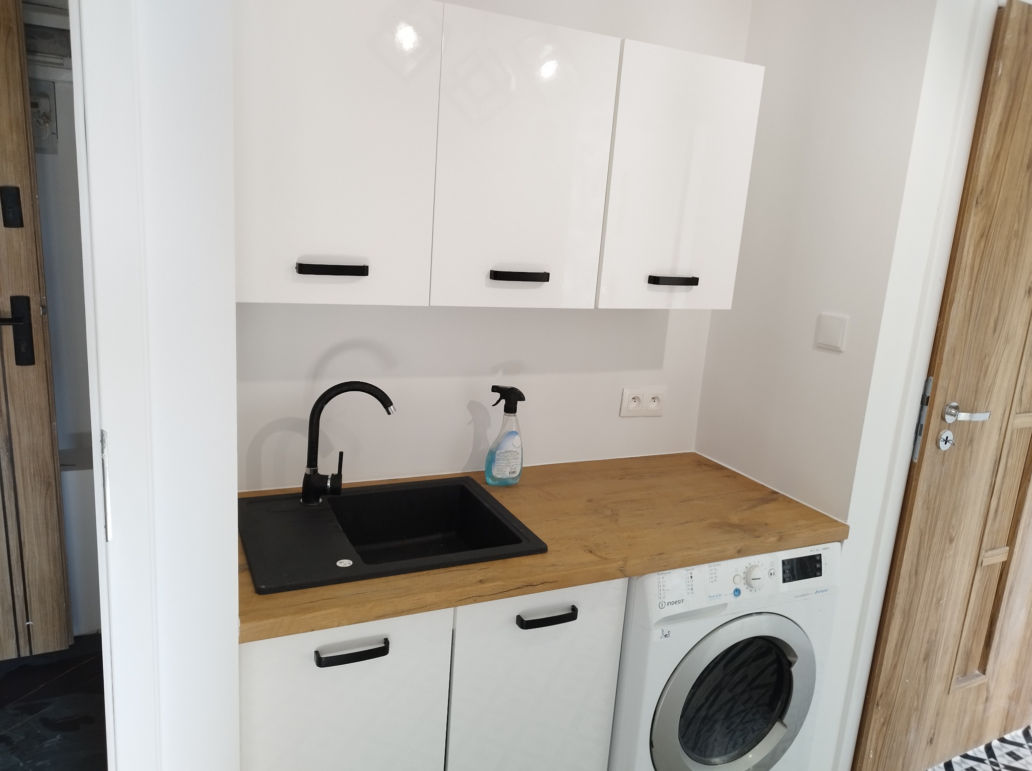 Nowoczesna aranżacja pralni z białymi szafkami, czarnym zlewem i baterią, drewnianym blatem oraz pralką Indesit. Na blacie stoi butelka płynu do czyszczenia.