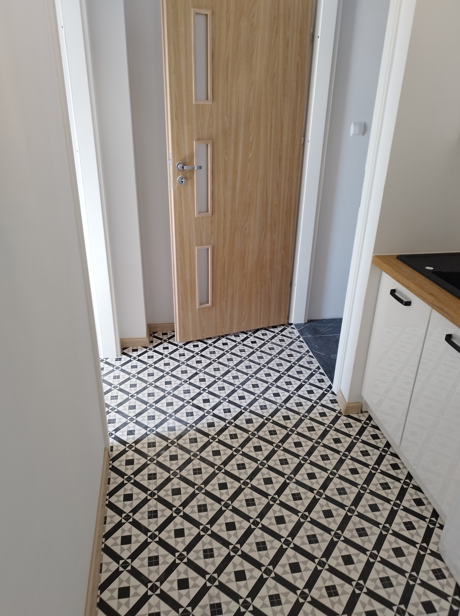 Ułożona czarno-biała podłoga w romby w przedpokoju z drewnianą ościeżnicą drzwi wewnętrznych i fragmentem białej zabudowy kuchennej.