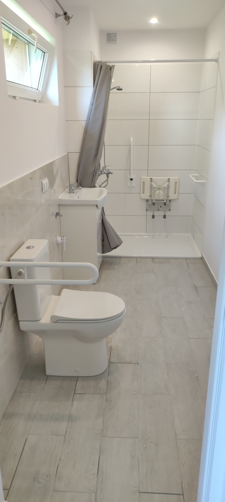 Jasna łazienka dostosowana dla osób niepełnosprawnych z toaletą z poręczami, umywalką z zasłoną, kabiną prysznicową i składanym siedziskiem pod prysznicem.
