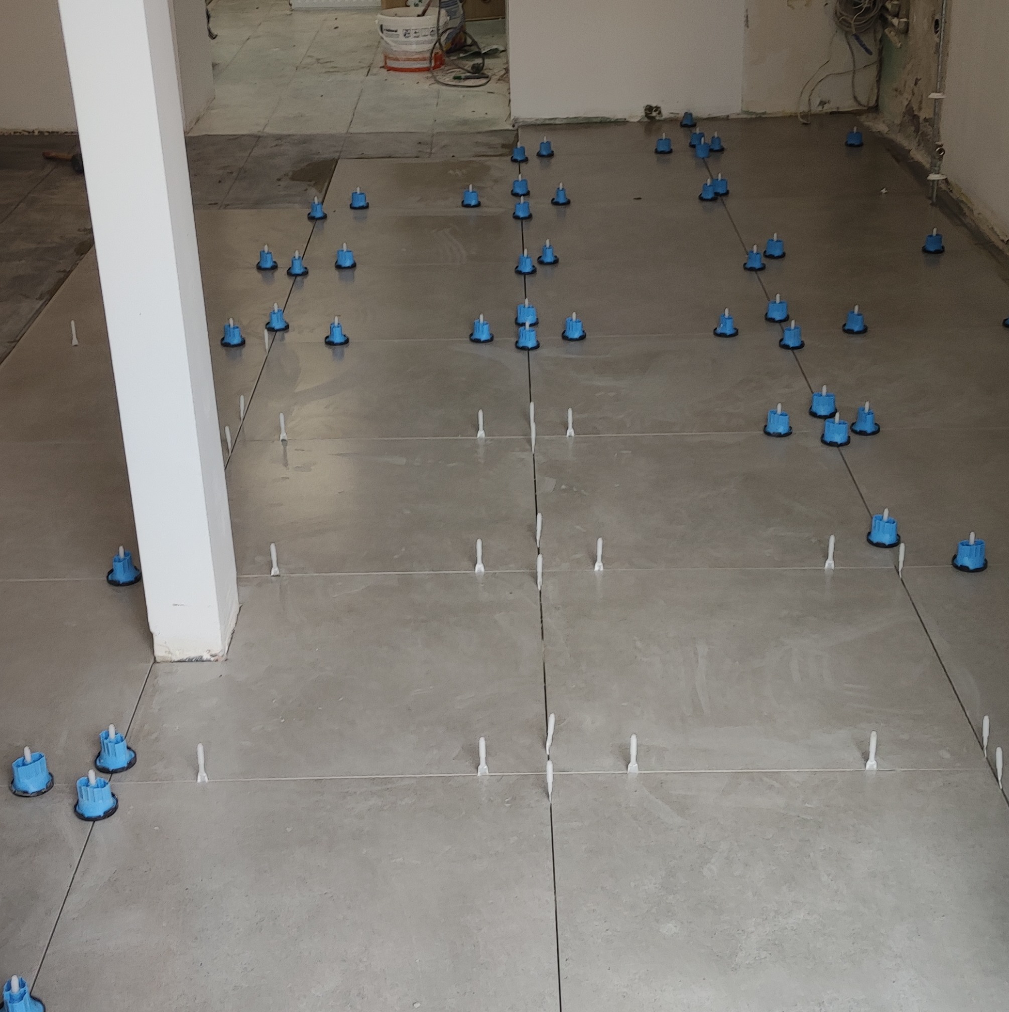 Świeżo ułożone, szare płytki podłogowe z niebieskimi klipsami poziomującymi i białymi krzyżykami dystansującymi spoiny, widoczne niedoskonałości ścian i słup nośny.