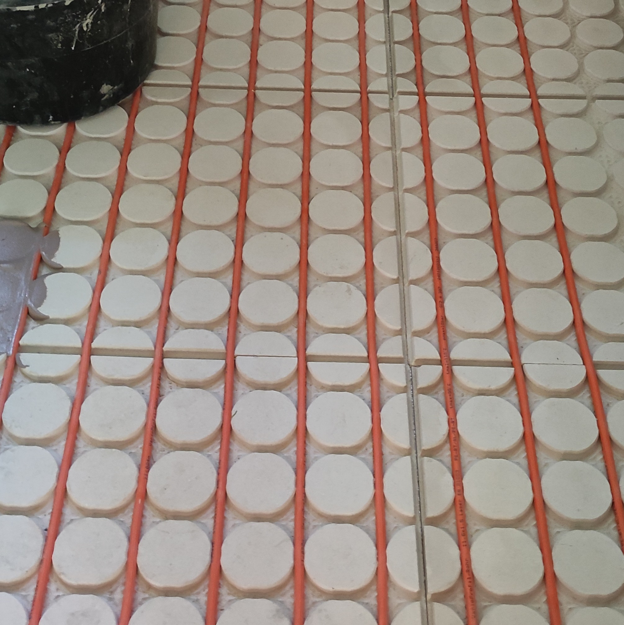 System ogrzewania podłogowego z rurami w kolorze pomarańczowym i białymi okrągłymi płytkami, widoczny fragment wiaderka w lewym górnym rogu.