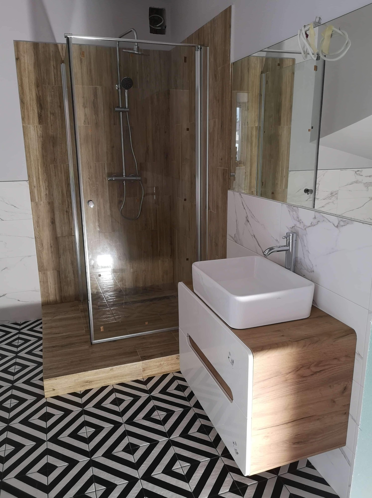 Nowoczesna łazienka z kabiną prysznicową wyłożoną płytkami imitującymi drewno, białą umywalką nablatową na szafce z drewnianym frontem i geometryczną posadzką w czarno-białe wzory.