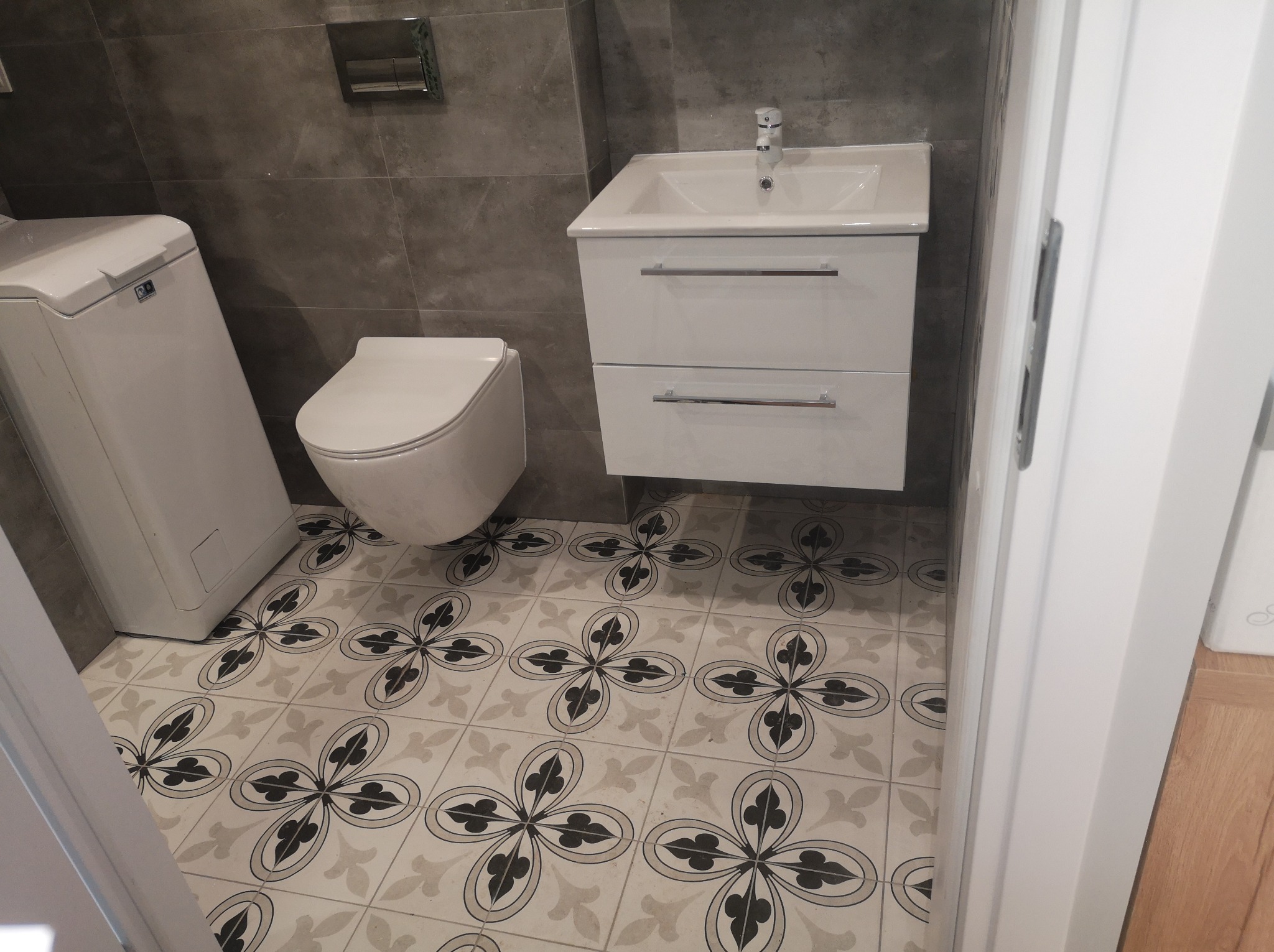 Nowoczesna łazienka z szarymi płytkami ściennymi, białą umywalką z szafką, podwieszaną toaletą i pralką, charakteryzująca się wzorzystą podłogą w kwiaty.