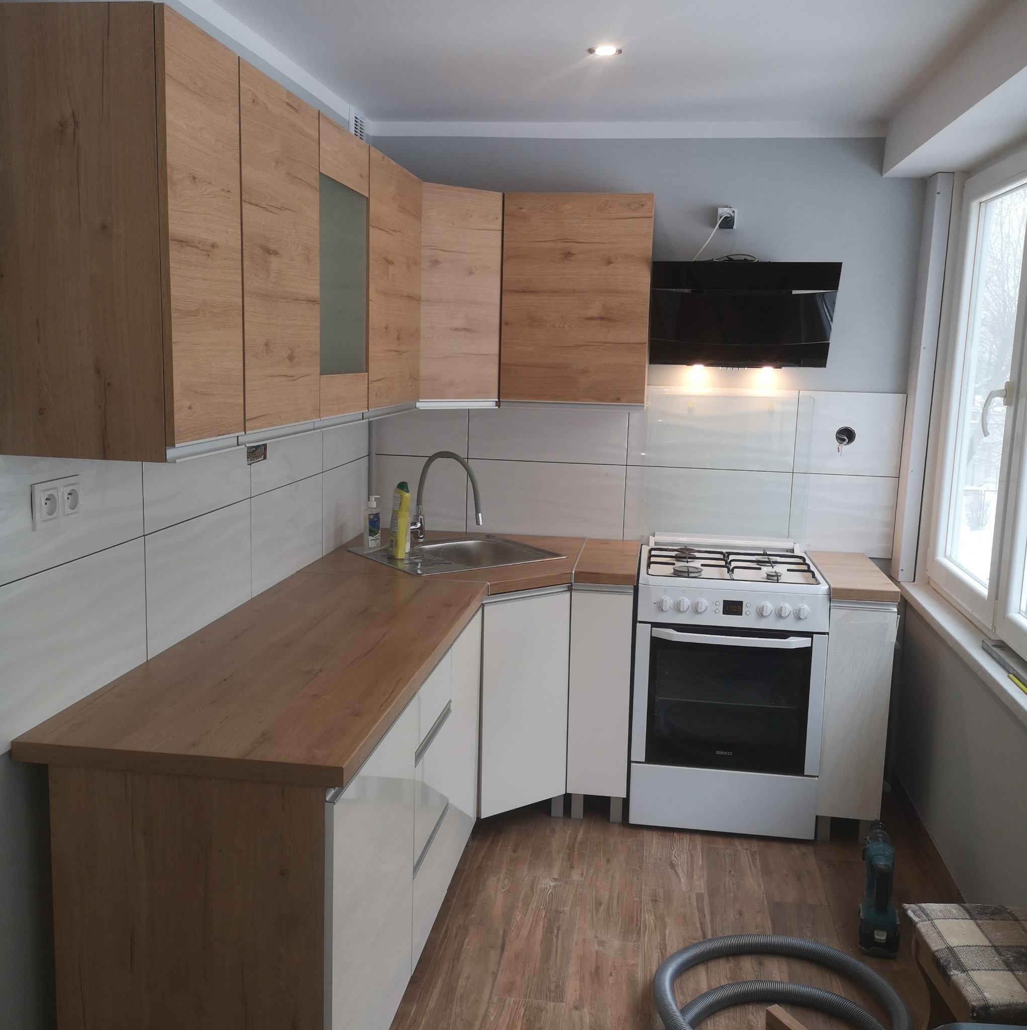 Nowoczesna kuchnia z drewnianymi szafkami wiszącymi i białymi szafkami dolnymi, białe płytki na ścianie, kuchenka gazowa i okap, widoczne narzędzia i odkurzacz na podłodze.