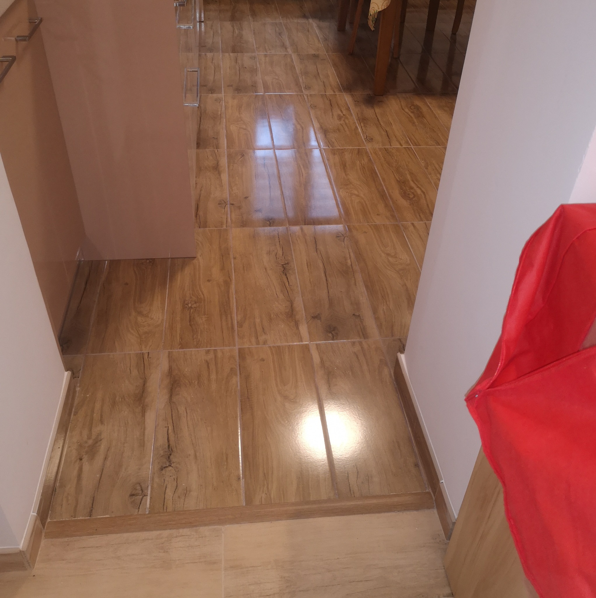 Ułożona podłoga z płytek imitujących drewno, widoczne fugi i listwy przypodłogowe, fragment mebla i czerwonej tkaniny w rogu kadru.