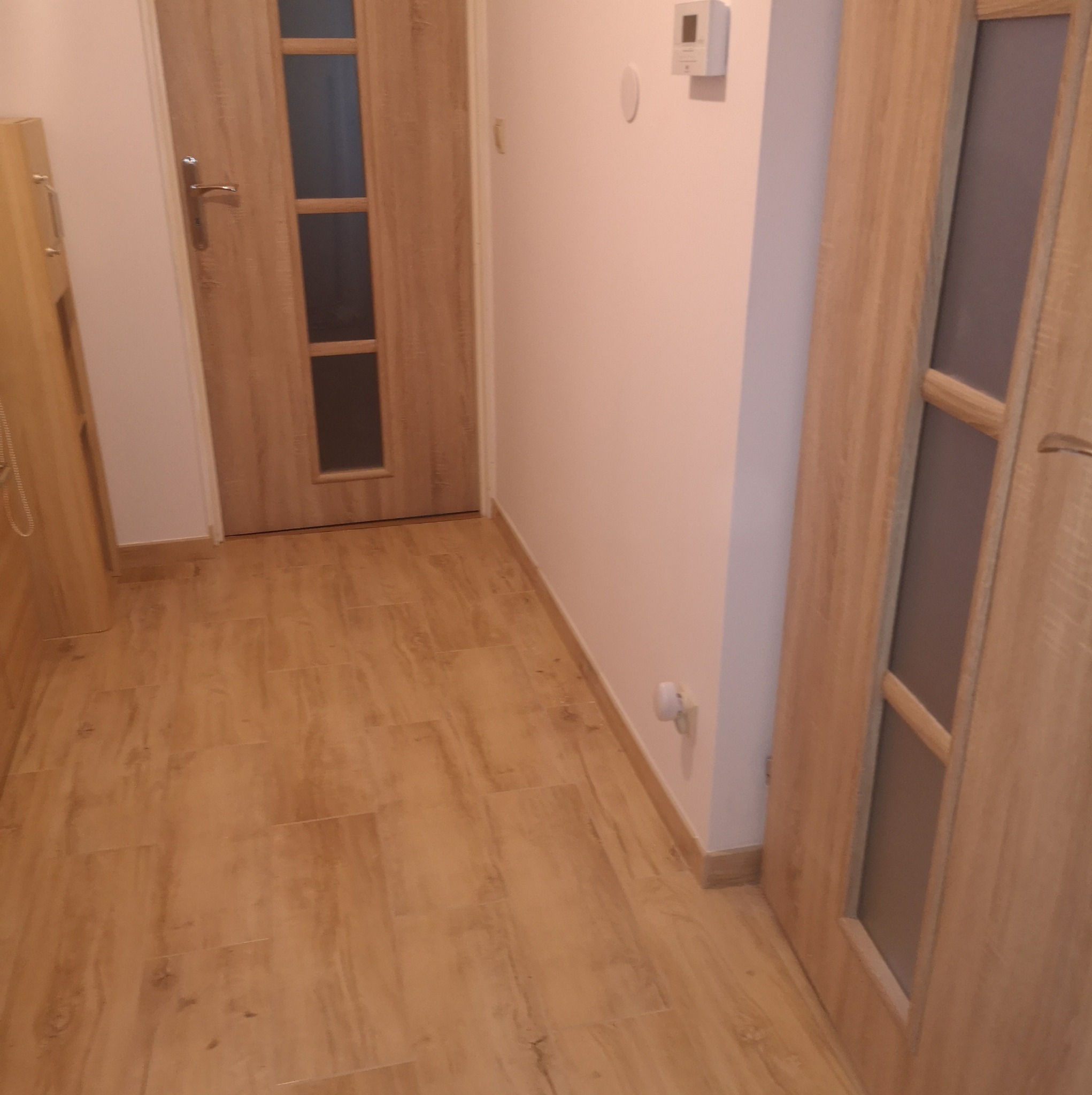 Korytarz wykończony płytkami imitującymi drewno, z widocznymi dwiema drewnianymi drzwiami z przeszkleniami i białymi ścianami z zamontowanym panelem sterowania ogrzewaniem.