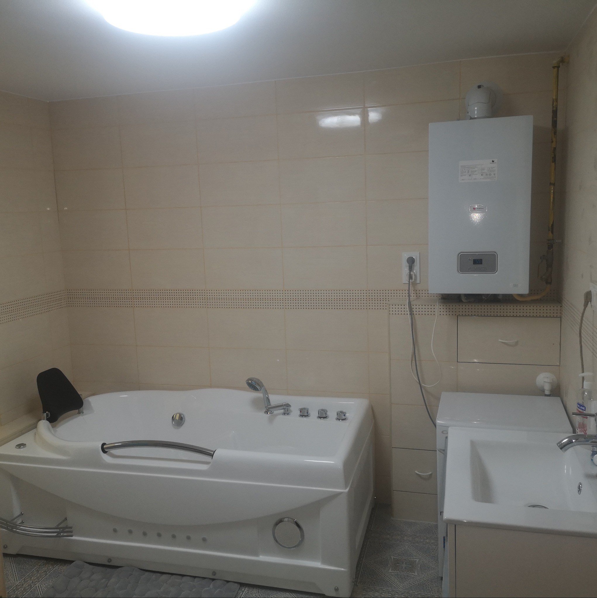 Wykończona łazienka z wanną z hydromasażem, umywalką z szafką i piecem gazowym na ścianie, kremowe płytki z dekoracyjnym pasem, szara mata na podłodze.