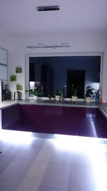 Nowoczesna kuchnia z fioletowymi szafkami, oświetleniem LED pod szafkami i lampą sufitową z okrągłymi elementami, widok na okno z roślinami na parapecie