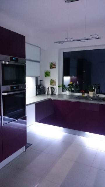 Nowoczesna kuchnia z fioletowymi szafkami, oświetleniem LED pod szafkami i srebrnymi akcentami, widok na okno z roślinami i akcesoriami kuchennymi.