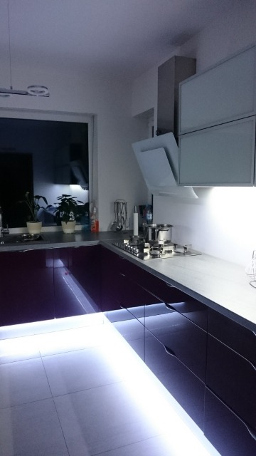 Nowoczesna kuchnia z burgundowymi szafkami, podświetleniem LED pod blatem i białymi szafkami wiszącymi oraz oknem z widokiem na ogród.