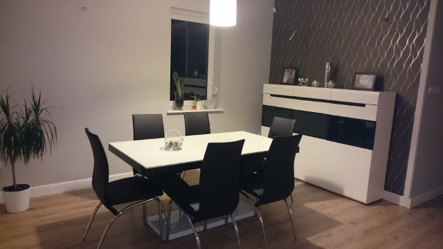 Jasna jadalnia z białym stołem, czarnymi krzesłami i białą komodą na tle wzorzystej tapety.