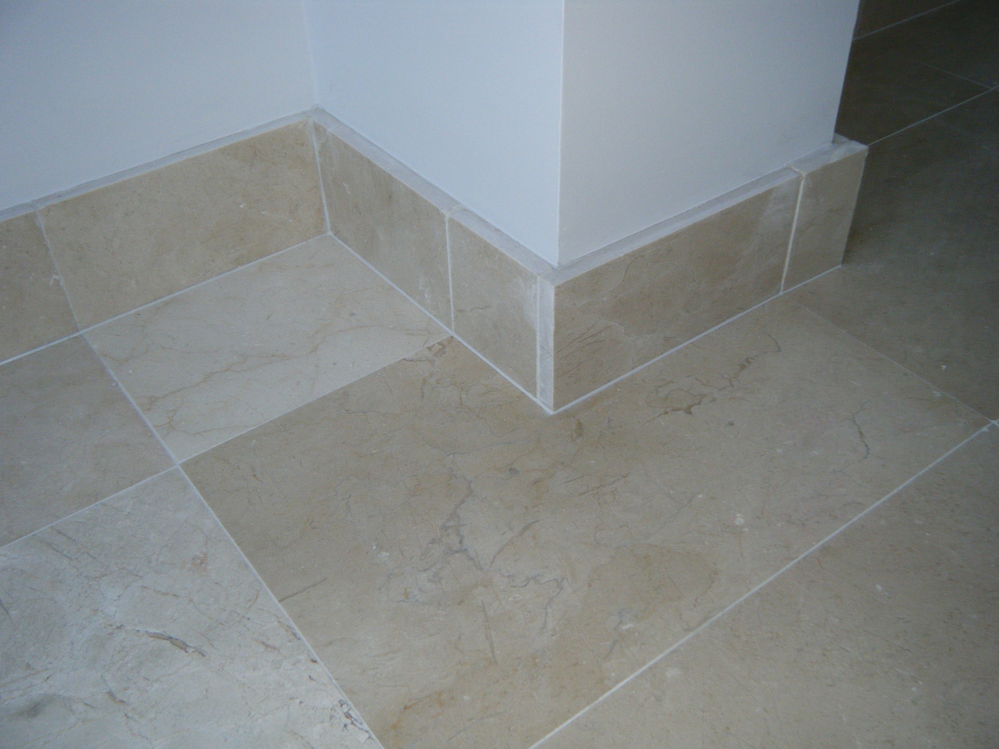 Wykończona podłoga z beżowych, marmurkowych płytek ceramicznych z listwami przypodłogowymi w narożniku pomieszczenia.