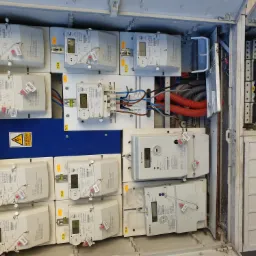 Otwarta skrzynka elektryczna z wieloma licznikami energii elektrycznej, widoczne kable i przewody w peszlach, tabliczka ostrzegawcza o zagrożeniu porażeniem elektrycznym.