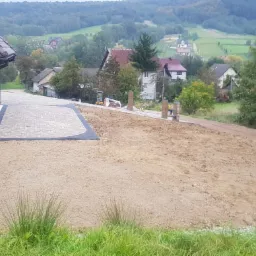 Układanie kostki brukowej na podjeździe nowego domu z widokiem na pagórkowaty krajobraz, częściowo przygotowany teren pod ogród.