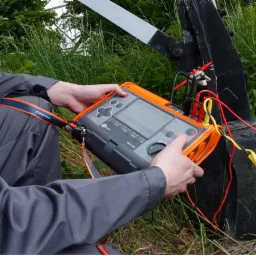 Elektryk w szarym kombinezonie wykonuje pomiary elektryczne przy użyciu miernika wielofunkcyjnego MIC-250 na słupie bramy wjazdowej, widoczne kable pomiarowe.