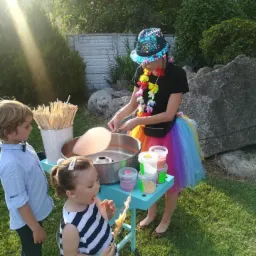 Kobieta w cekinowym kapeluszu i tęczowej spódnicy robi watę cukrową dla dzieci na zielonej trawie w słoneczny dzień.